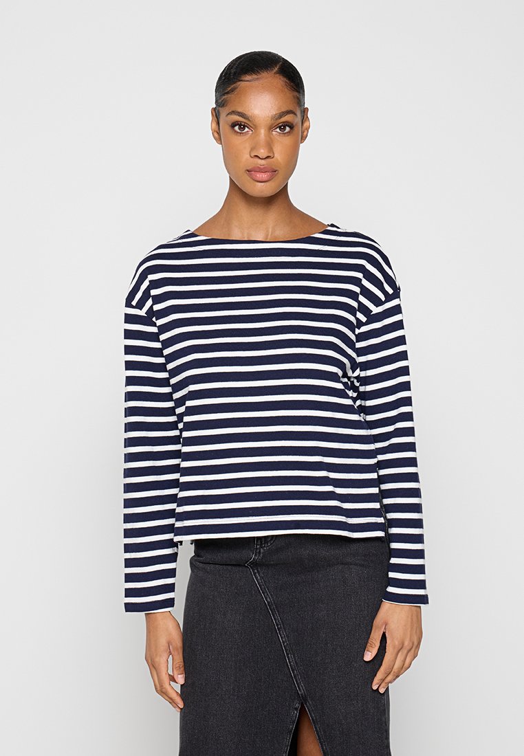 Frau trägt ein marineblau-weiß horizontal gestreiftes Langarmshirt und einen dunkelgrauen, hoch taillierten Rock mit Frontschlitz.