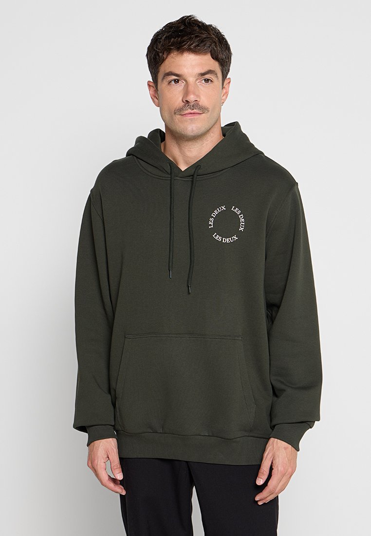 Dunkelgrüner Kapuzenpullover aus weichem Stoff, mit einer Vordertasche, Kordelzug und rundem weißen Logo auf der Brust. Entspannt geschnittenes Design.