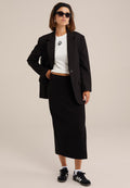 Schwarze taillierte Blazer über einem weißen Cropped-Top mit schwarzem Logo, kombiniert mit einem schwarzen Midi-Rock und schwarzen Sneakers mit weißen Streifen.