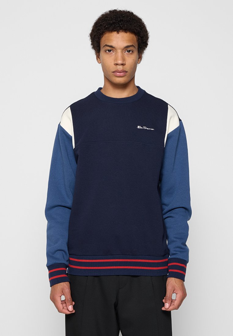 Junger Mann, der einen navyblauen und weißen Langarm-Sweatshirt mit rotgestreiften Bündchen und Saum trägt, steht vor einem schlichten hellen Hintergrund.