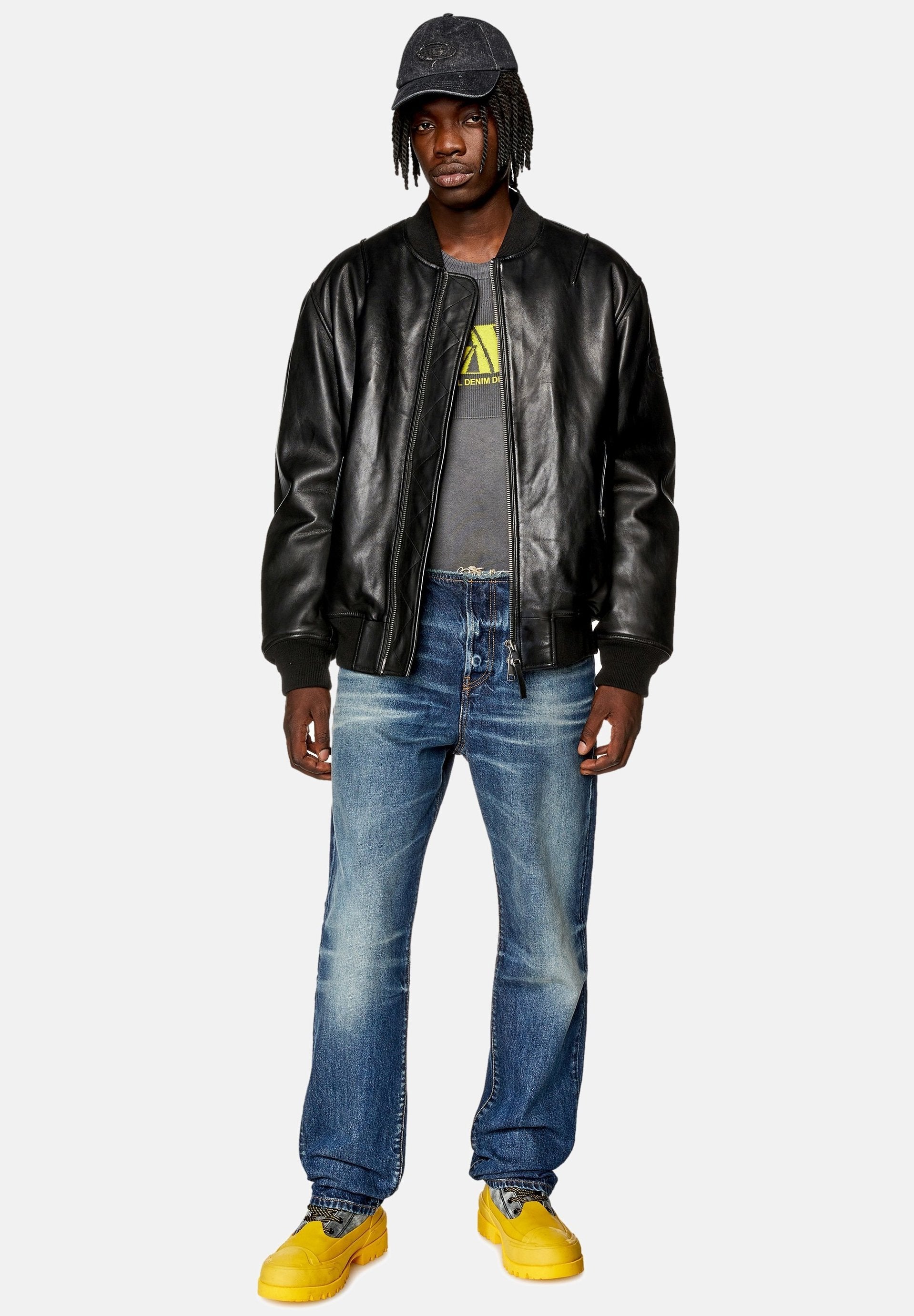 Schwarze Lederbomberjacke, graues Grafik-T-Shirt, blaue Denim-Jeans und gelbe Sneaker. Zubehör umfasst eine dunkle Kappe. Lässiges Outfit.