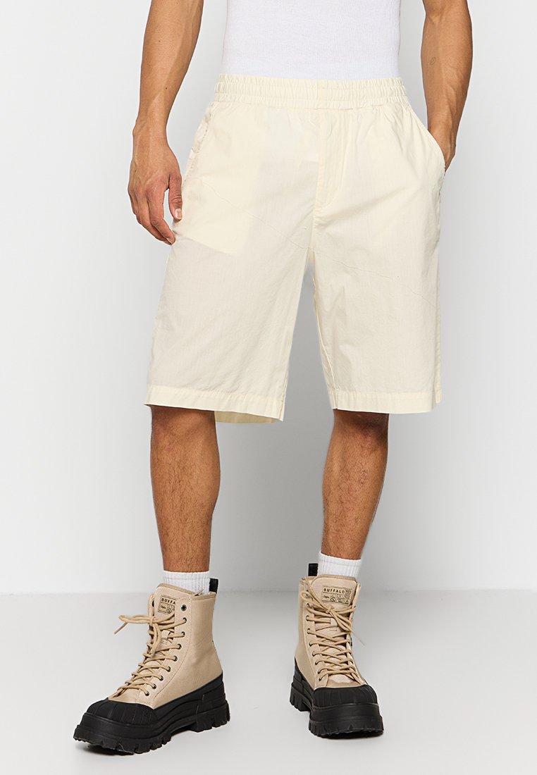 Mann in beigen, knielangen Shorts mit elastischem Bund, weißen Socken und beigen High-Top-Stiefeln mit schwarzen Sohlen.