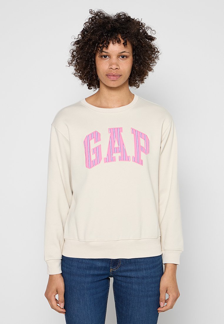 Frau mit lockigem Haar, die einen cremefarbenen Pullover mit rosa "GAP"-Schriftzug und blaue Jeans trägt, steht vor einem schlichten hellgrauen Hintergrund.