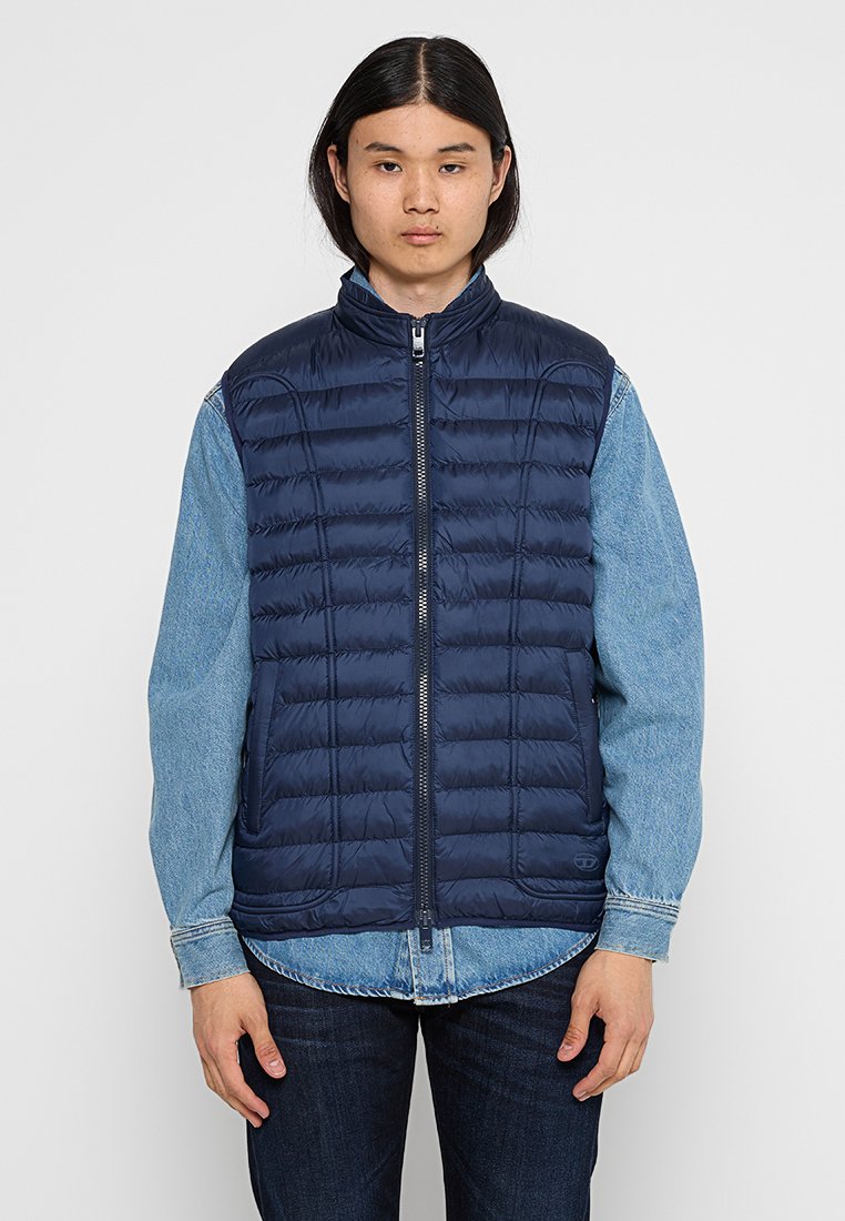 marineblaues Steppweste mit horizontalen Nähten und hohem Kragen, ausgestattet mit einem Frontreißverschluss und seitlichen Taschen, getragen über einem Jeanshemd.