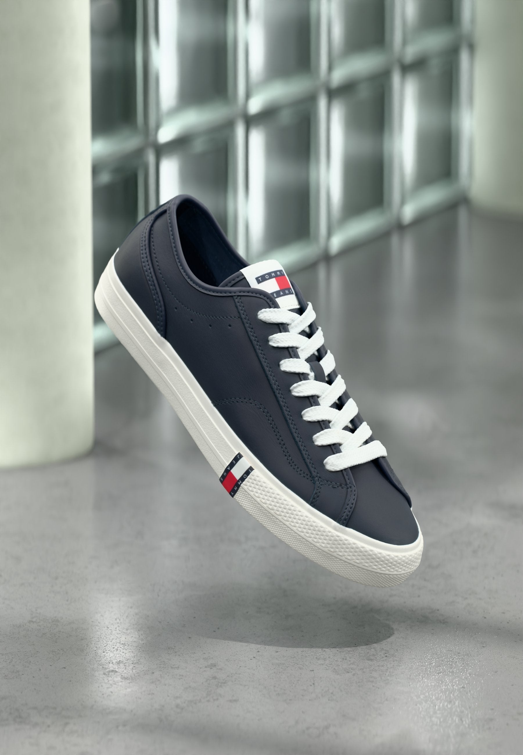 Marineblaue Sneaker mit weißen Schnürsenkeln und Sohle, mit Tommy Hilfiger Logo auf der Zunge und der Seite, schwebend über einem grauen Boden im Innenbereich.