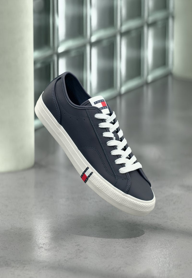 Marineblaue Sneaker mit weißen Schnürsenkeln und Sohle, mit Tommy Hilfiger Logo auf der Zunge und der Seite, schwebend über einem grauen Boden im Innenbereich.