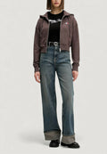 Braune Kapuzenjacke mit Reißverschluss und Taschen, kombiniert mit einem schwarzen, gerippten Shirt mit weißem Logo. Hoch taillierte blaue Jeans mit weitem Bein und kontrastierenden Bündchen.