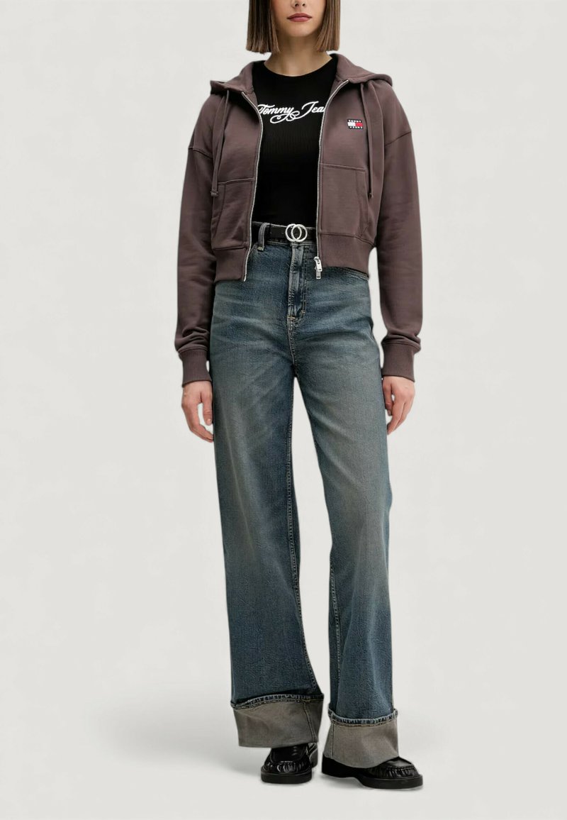 Braune Kapuzenjacke mit Reißverschluss und Taschen, kombiniert mit einem schwarzen, gerippten Shirt mit weißem Logo. Hoch taillierte blaue Jeans mit weitem Bein und kontrastierenden Bündchen.