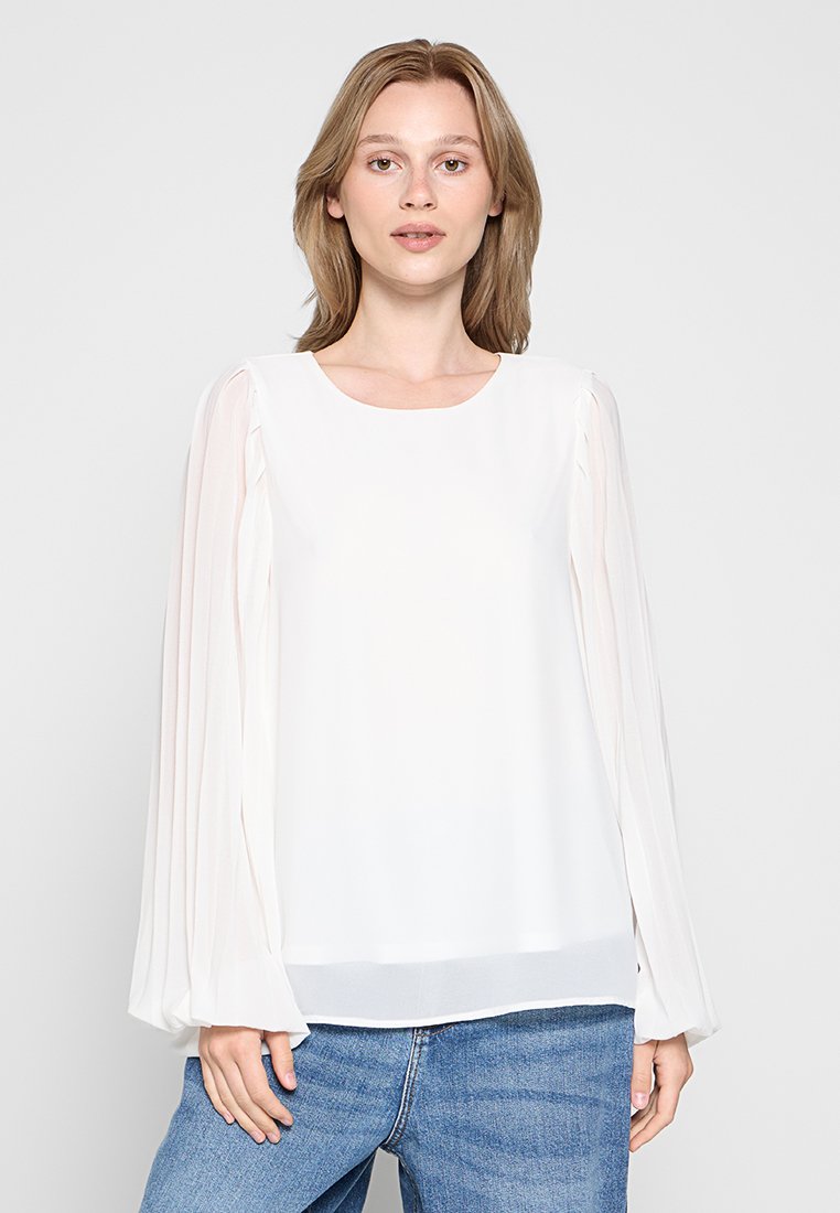 Weißes Blusenshirt mit transparenten, plissierten langen Ärmeln und rundem Halsausschnitt. Glatter Stoff mit lockerem Schnitt, kombiniert mit blauen Jeans.