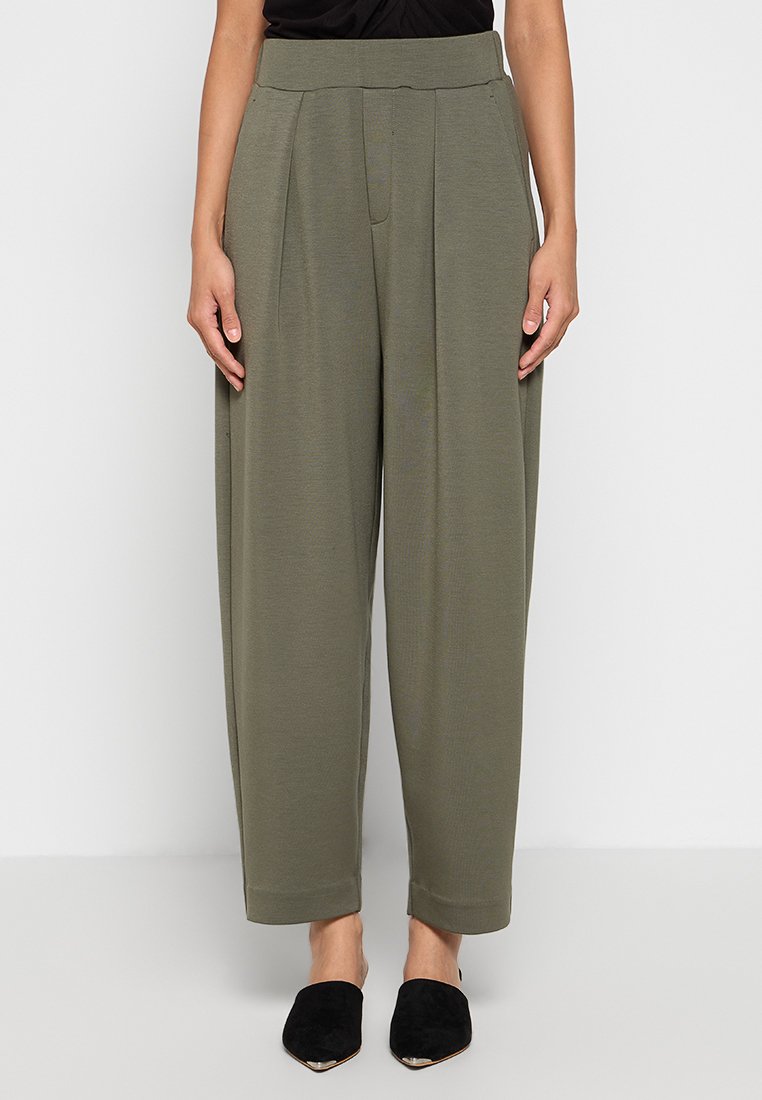 Olivgrüne, maßgeschneiderte Hose mit hohem Bund und Falten. Sanfte Textur, weiter Beinschnitt, gesäumte Säume, kombiniert mit schwarzen Slippern.