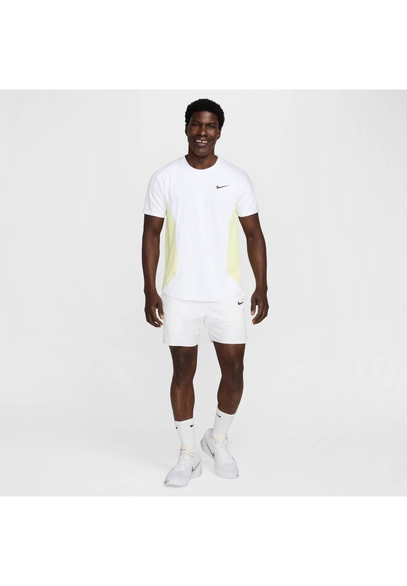 Männliches Sportoutfit mit einem weißen T-Shirt, das helle gelbe Seitenpanels und ein schwarzes Logo hat, ergänzt durch weiße Shorts und Socken.