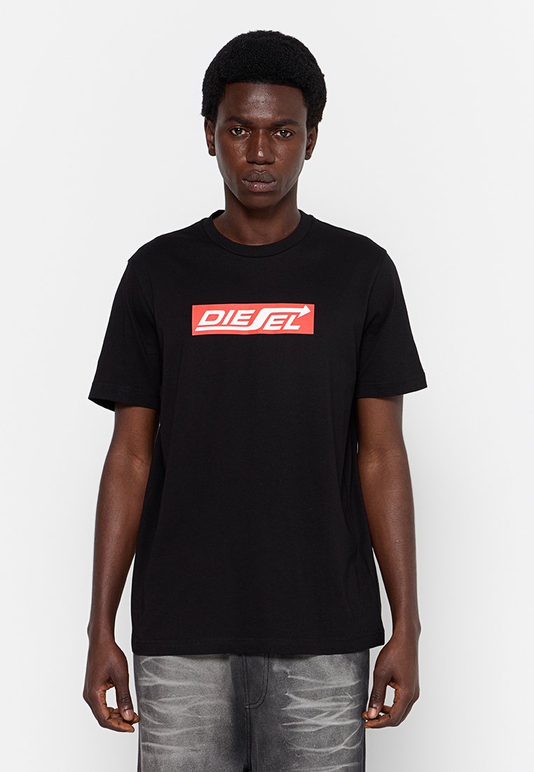 Junger Mann mit kurzen lockigen Haaren, der ein schwarzes Diesel-T-Shirt mit rotem Logo und grauen gemusterten Jeans trägt, vor einem schlichten weißen Hintergrund stehende.