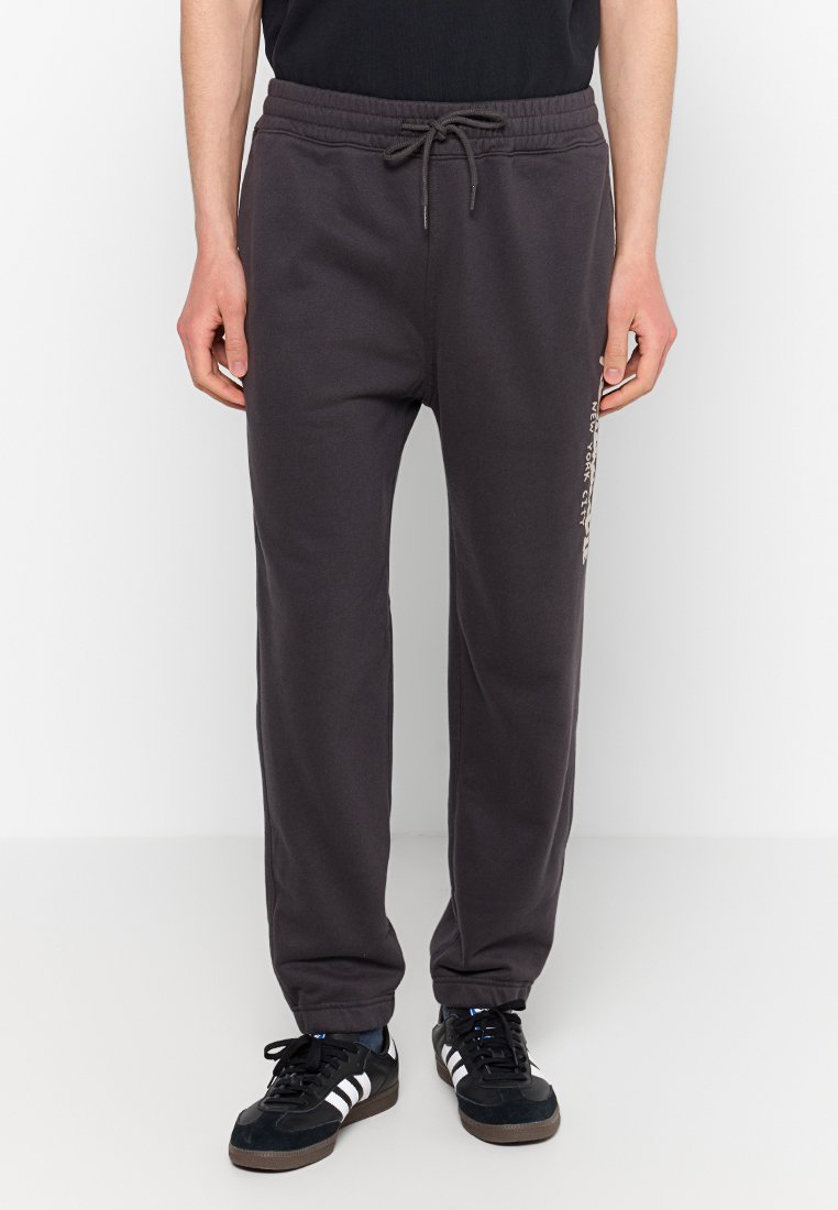 Mann in schwarzen Sweatpants mit Zugband und dem Schriftzug "New York City" an der Seite, sowie schwarzen Turnschuhen mit weißen Streifen, steht auf einem weißen Boden.
