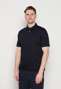 Mann steht vor einem einfarbigen Hintergrund, trägt ein schwarzes Poloshirt mit kurzen Ärmeln und dunkle Hose, blickt nach vorne mit neutralem Gesichtsausdruck.