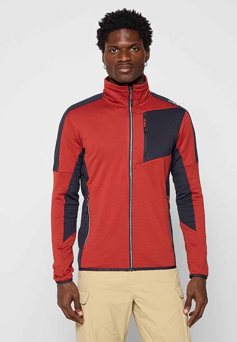 Mann in einer rot-schwarzen Jacke mit Reißverschluss und mit Reißverschlusstaschen, beigen Hosen, der vor einem einfachen hellen Hintergrund steht.