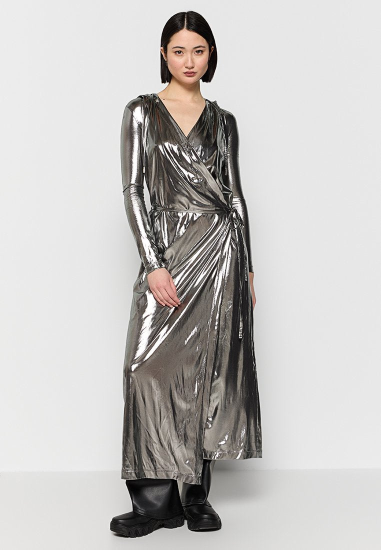 Frau mit glattem schwarzen Haar, die ein glänzendes, metallisches Wickelkleid mit langen Ärmeln und schwarze klobige Stiefel trägt, stehend vor einem schlichten Hintergrund.