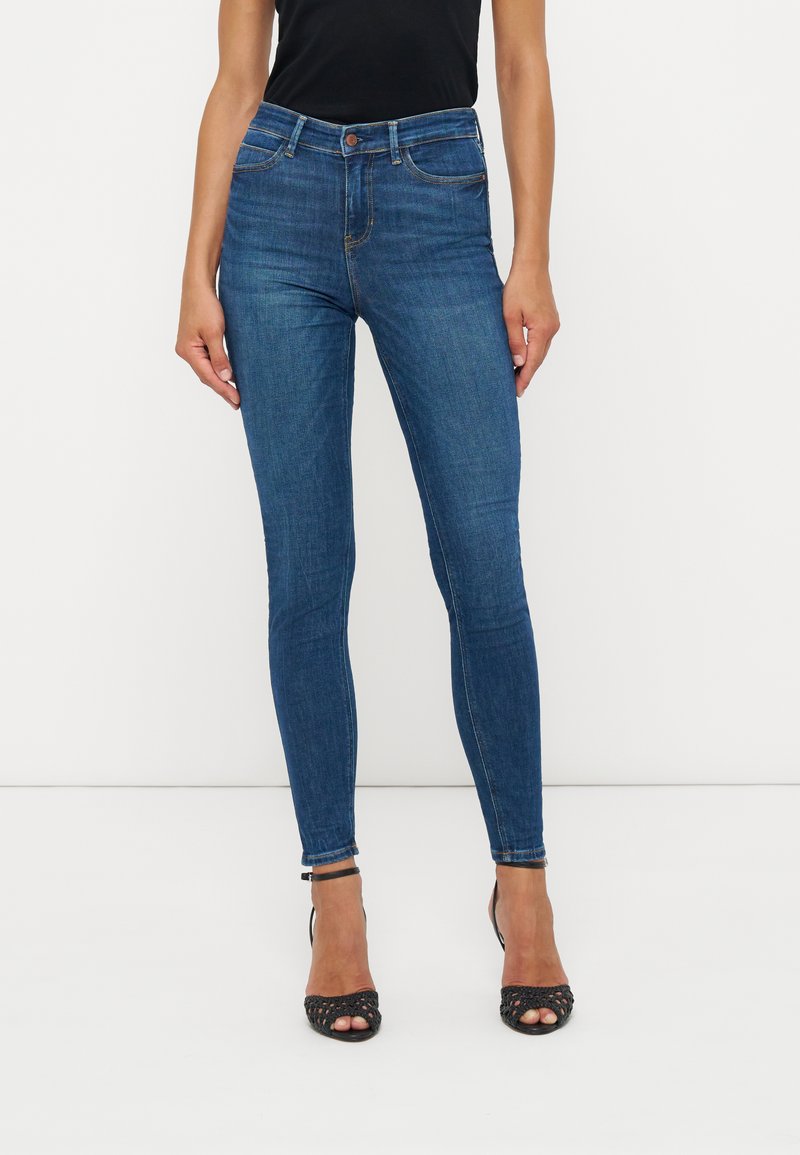 Frau trägt eng anliegende blaue Jeans und schwarze offene High-Heel-Sandalen, steht vor einem weißen Hintergrund.