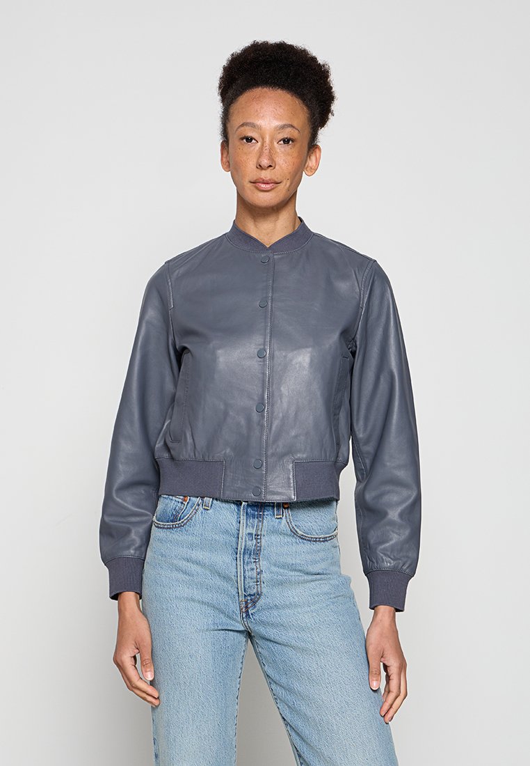 Frau mit lockigem Haar, die eine graue, geknöpfte Leder-Bomberjacke und hellblaue, hoch taillierte Jeans trägt und vor einem einfarbigen Hintergrund steht.