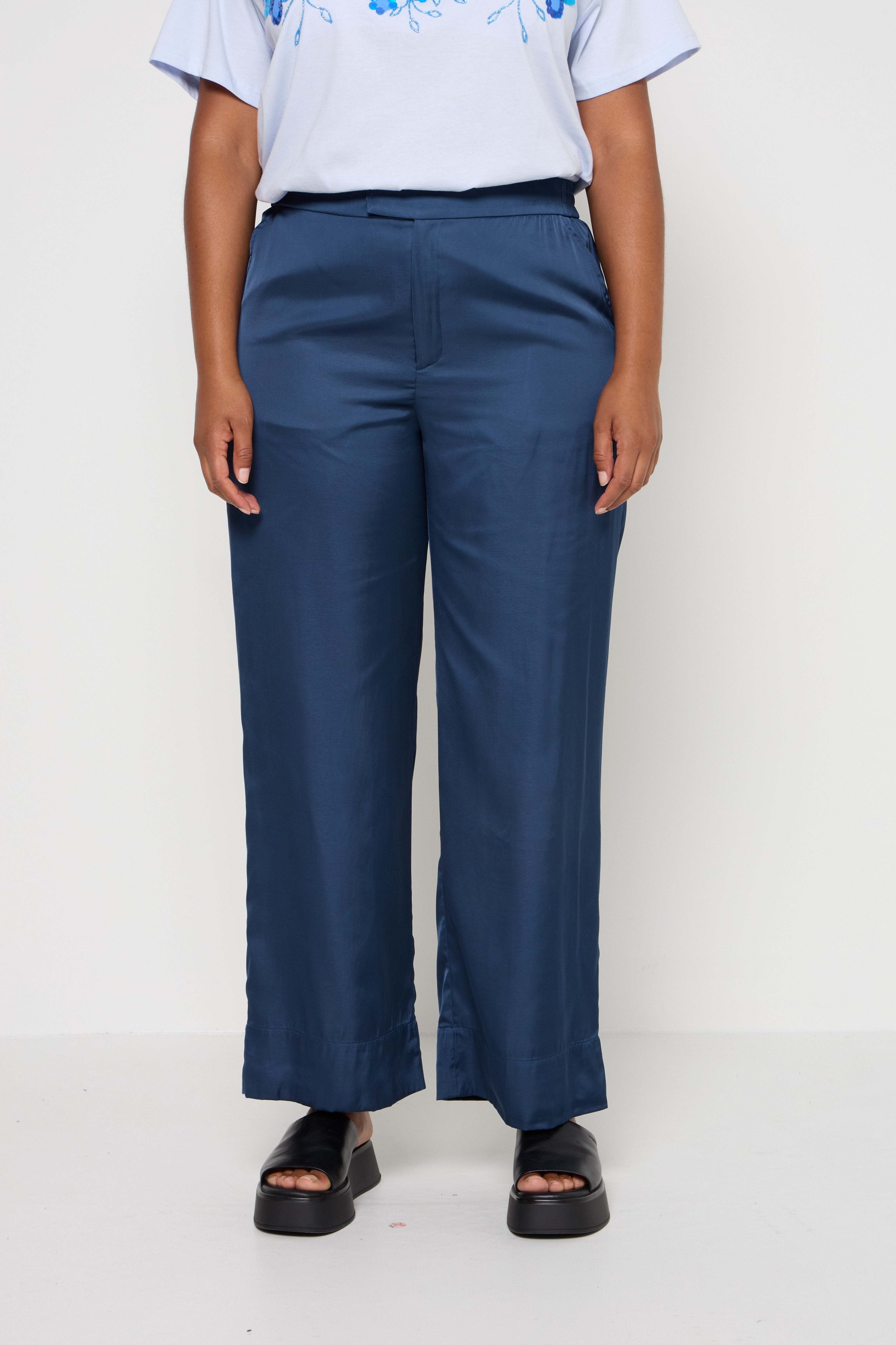Satinweite Hosen in Marineblau mit glatter Textur, ausgestattet mit einem Bund und dezenten seitlichen Details, kombiniert mit schwarzen Plateausandalen.