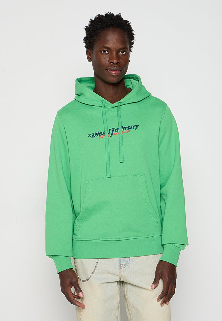 Junger Mann mit einem leuchtend grünen Hoodie mit dem Text "Diesel Industry Denim Division" und hellbeigen Jeans mit einer silbernen Kette.