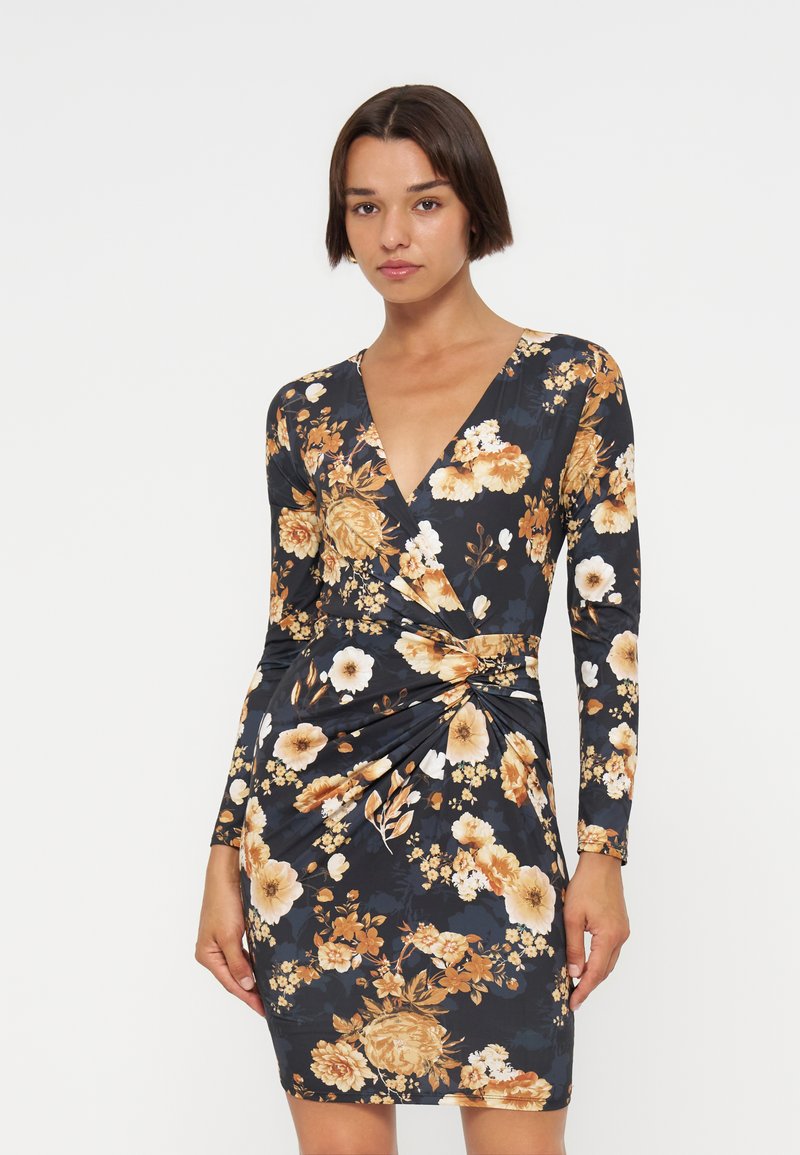 Frau mit kurzen dunklen Haaren, die ein langärmliges, tief ausgeschnittenes schwarzes Kleid mit orange-beigen Blumenmustern trägt.