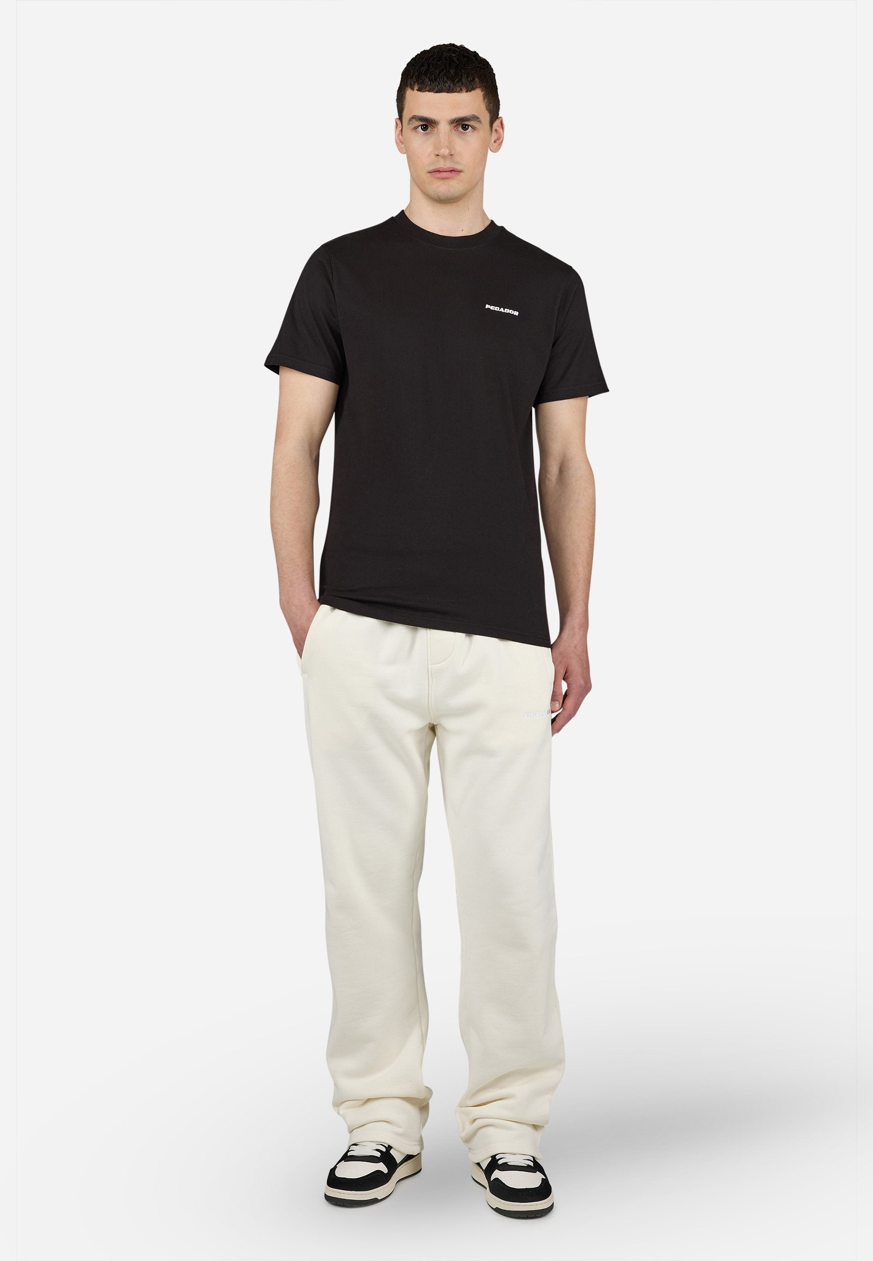 Junger Mann steht mit Händen in den Taschen, trägt schwarzes kurzärmeliges Hemd, hellbeige Hose und schwarz-weiße Sneakers vor weißem Hintergrund.
