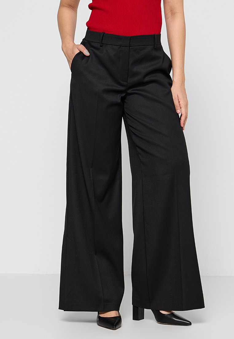 Schwarze, weit geschnittene Hosen mit glatter Textur, zwei seitlichen Taschen und taillierter Passform, kombiniert mit schwarzen spitzen High Heels.