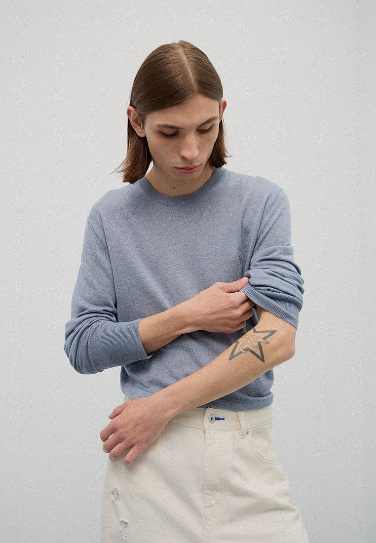 Himmelblaues langärmliges Sweatshirt aus strukturiertem Strick, Rundhalsausschnitt, mit eng anliegenden Ärmeln. Das Modell posiert mit cremefarbener, ausgebleichter Jeans und zeigt ein Tattoo.