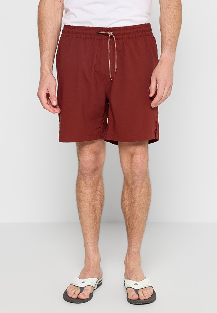Mann in burgunderfarbenen Z drawstring-Shorts, weißem T-Shirt und weißen Flip-Flops steht vor einem schlichten Hintergrund.