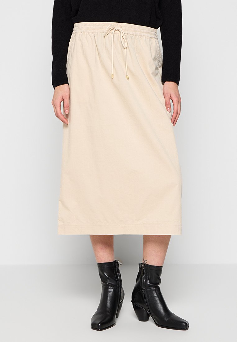 Beige, knielanger Rock aus Baumwolle mit elastischem Bund und Kordelzug. Kombiniert mit schwarzen, mit Reißverschluss versehenen Stiefeletten.