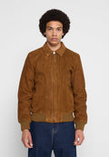 Junger Mann mit lockigem Haar, der eine braune Wildlederjacke mit Reißverschluss, gerippten Bündchen und Kragen sowie blaue Jeans trägt, steht vor einem schlichten Hintergrund.