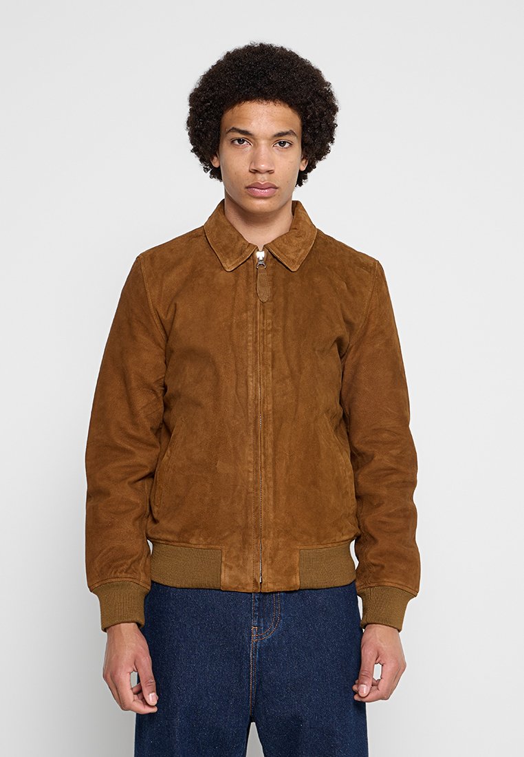Junger Mann mit lockigem Haar, der eine braune Wildlederjacke mit Reißverschluss, gerippten Bündchen und Kragen sowie blaue Jeans trägt, steht vor einem schlichten Hintergrund.