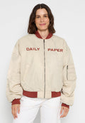 Beige Bomberjacke mit gerippten kastanienbraunen Bündchen und Kragen, mit "DAILY PAPER" in Rot auf der Brust bestickt und einer Reißverschlusstasche am Ärmel.