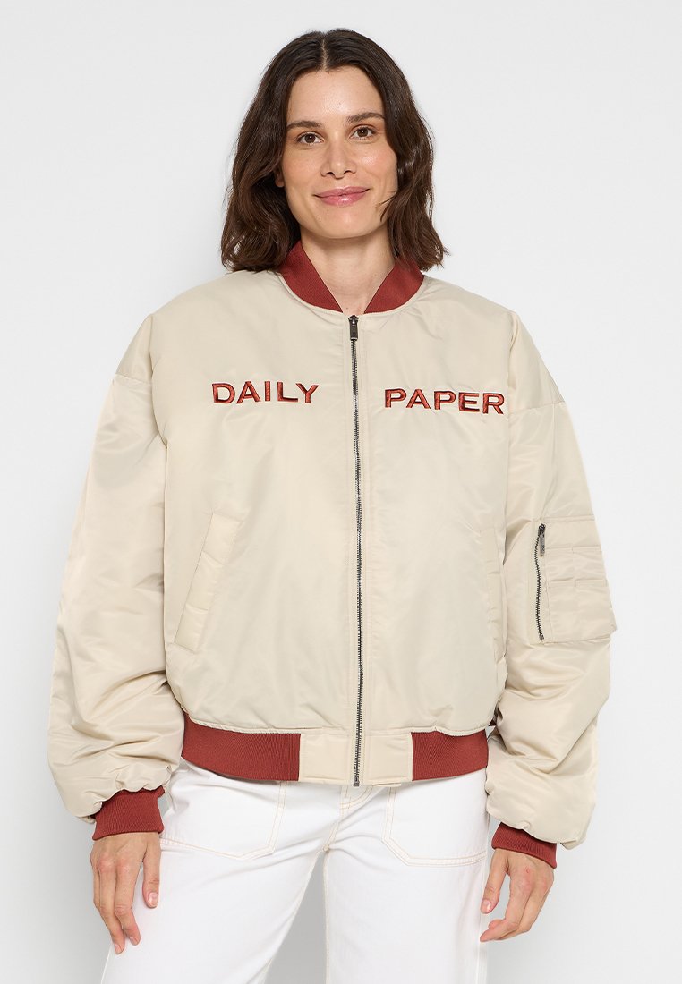 Beige Bomberjacke mit gerippten kastanienbraunen Bündchen und Kragen, mit "DAILY PAPER" in Rot auf der Brust bestickt und einer Reißverschlusstasche am Ärmel.
