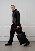 Schwarzer Hoodie mit Tasche vorne, rot-schwarz kariertes Hemd um die Taille gebunden, schwarze Hose und schwarze Tragetasche. Flache Schuhe.