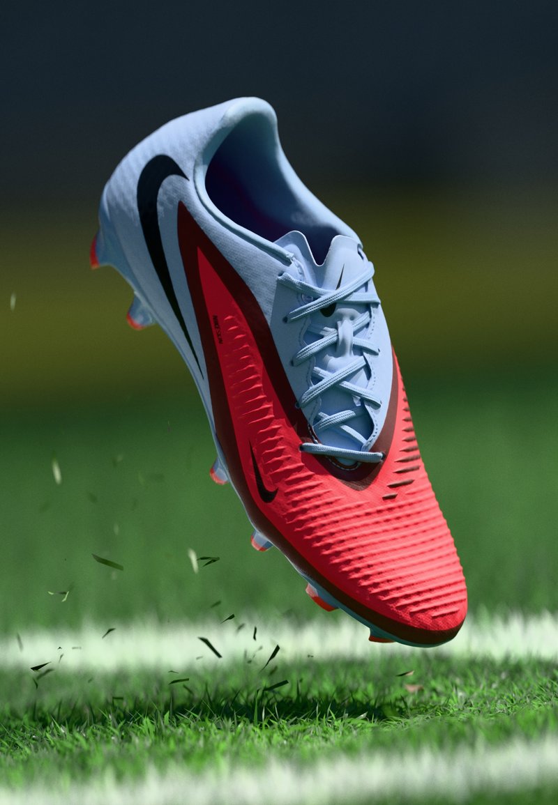 Rote und graue Fußballschuhe mit strukturierten Oberflächen, schwarzem Swoosh und hellblauen Schnürsenkeln, über Gras mit Rasenabfällen erhoben.
