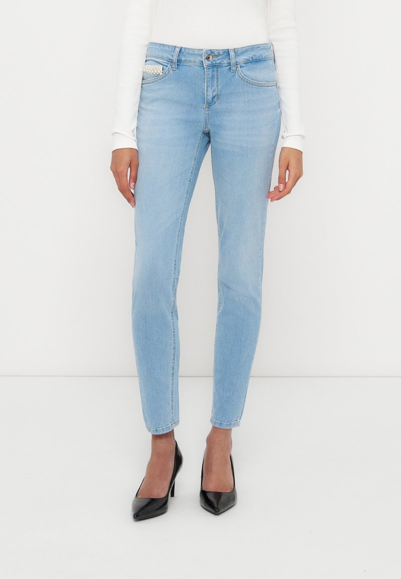 Frau trägt hellblaue, eng anliegende Jeans mit Perlendetail an der Vordertasche, weißes langärmeliges Oberteil und schwarze High Heels.
