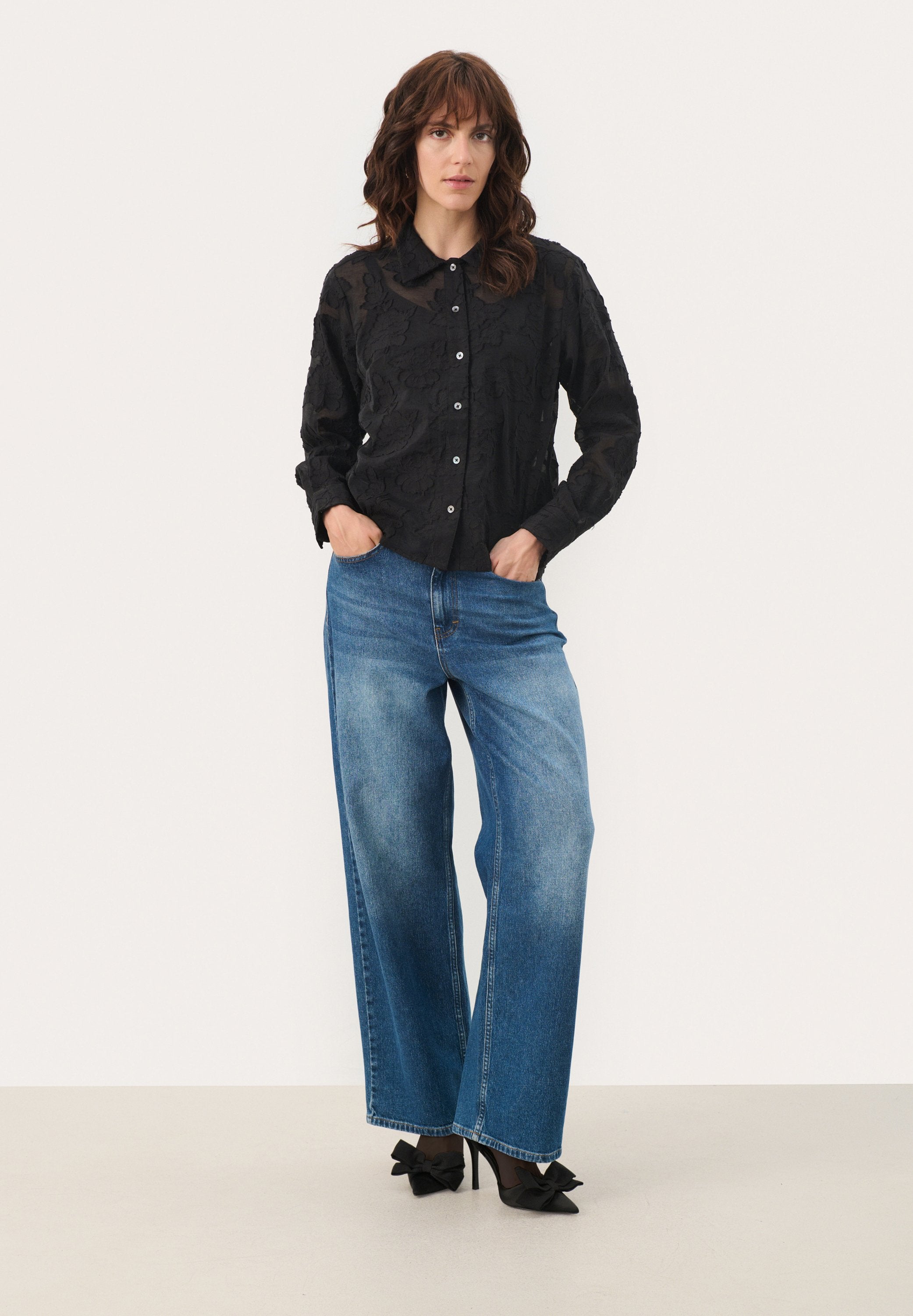 Schwarzes, blumenmusterndes Hemd mit Knöpfen und Kragen, kombiniert mit weiten, hellblauen Denim-Jeans und schwarzen High Heels mit Schleifen.
