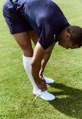 Weiße Fußballschuhe mit strukturierten Details, getragen mit weißen Socken und dunklen Sportshorts, während eine Person ihre Schnürsenkel auf dem Gras bindet.