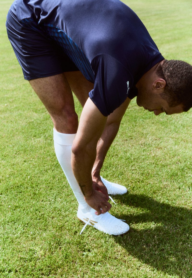 Weiße Fußballschuhe mit strukturierten Details, getragen mit weißen Socken und dunklen Sportshorts, während eine Person ihre Schnürsenkel auf dem Gras bindet.