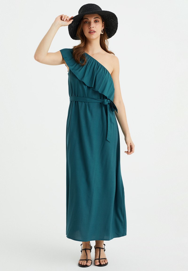 Türkisfarbenes Maxikleid mit einem einseitigen Design, Rüschendetails und einem Taillengürtel, kombiniert mit einem schwarzen Hut mit breiter Krempe und Riemchensandalen.