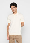 Mann mit lockigem Haar, der ein cremefarbenes Kurzarm-Polohemd und beige Hosen trägt, steht mit einer Hand hinter dem Rücken.