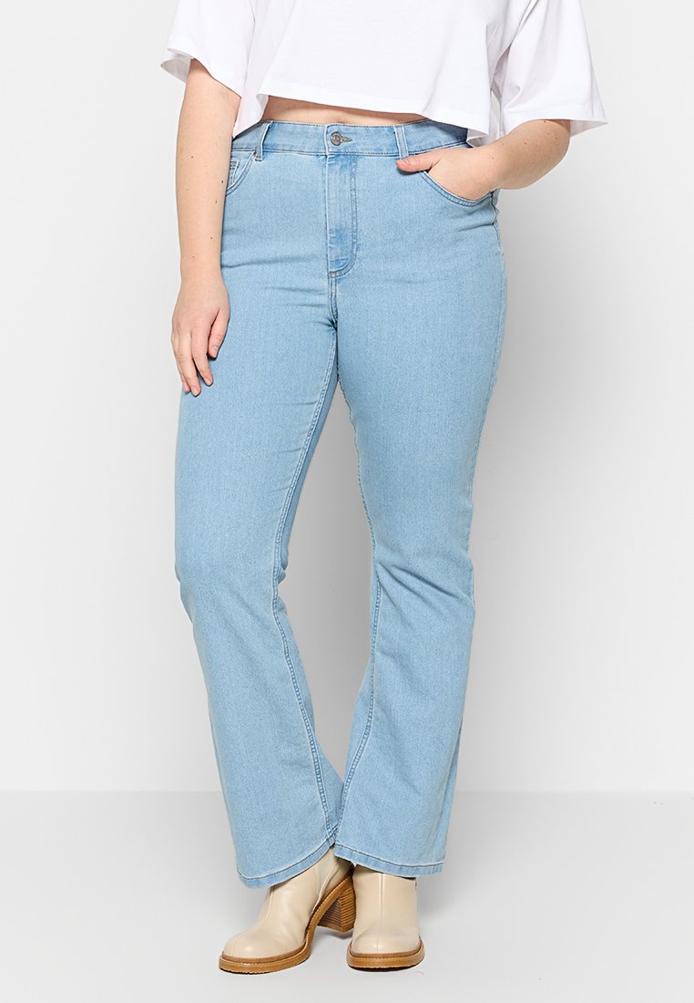Hellblaue Jeans im Schlagstil mit hoher Taille, ausgestattet mit einem Reißverschluss, seitlichen Taschen und dezenten Nähdetails. Kombiniert mit beigen Stiefeletten.