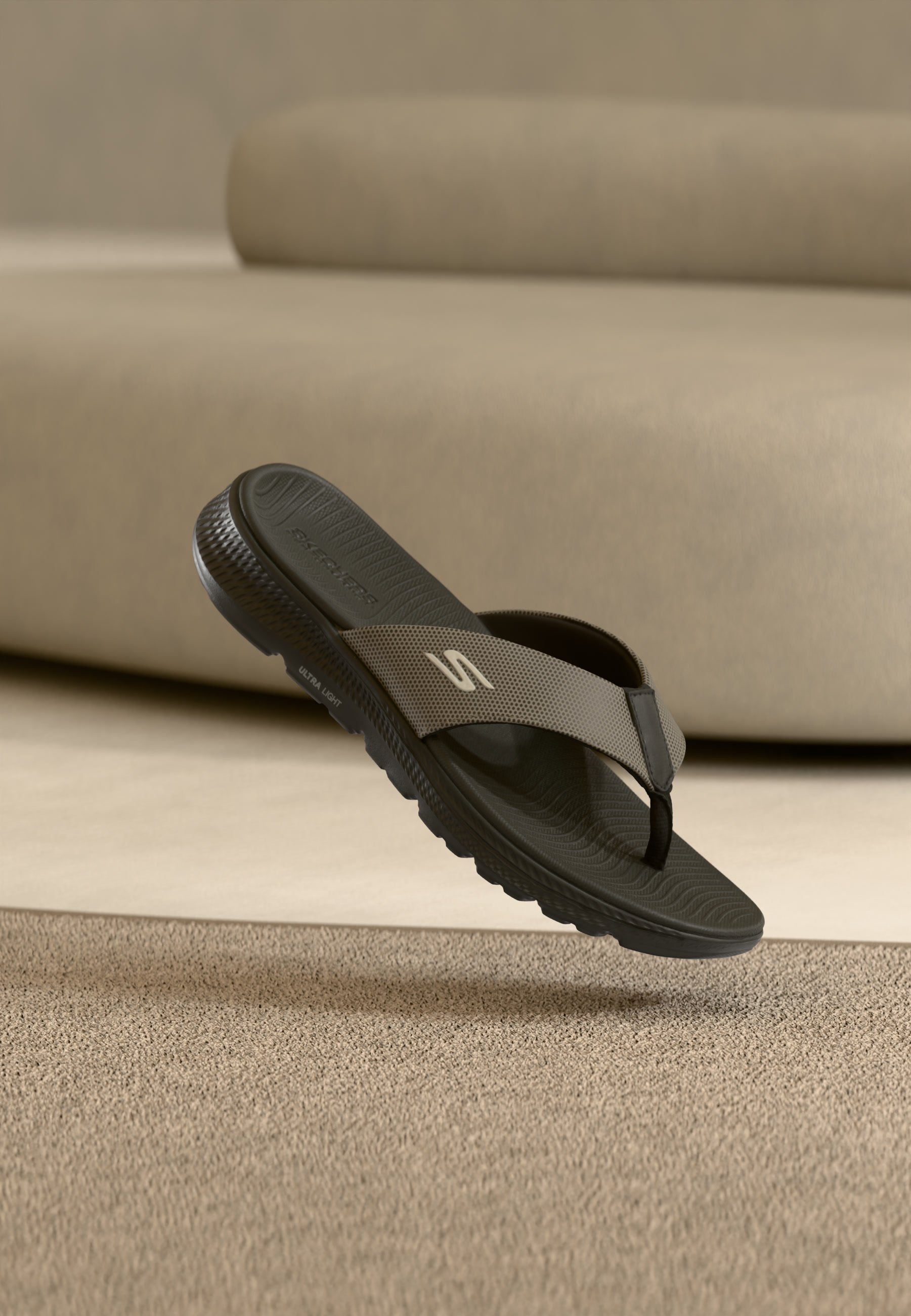 Schwarze und graue Skechers Zehensteg-Sandale mit strukturierter Sohle, die über einem beigen Teppich vor einem beigen Sofa schwebt.