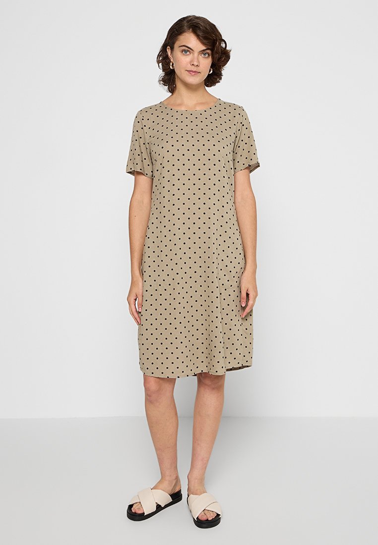 Beige Kleid bis zum Knie mit kurzen Ärmeln und schwarzen Tupfen; runder Ausschnitt; lockere Passform; kombiniert mit hellen, lässigen Sandalen.