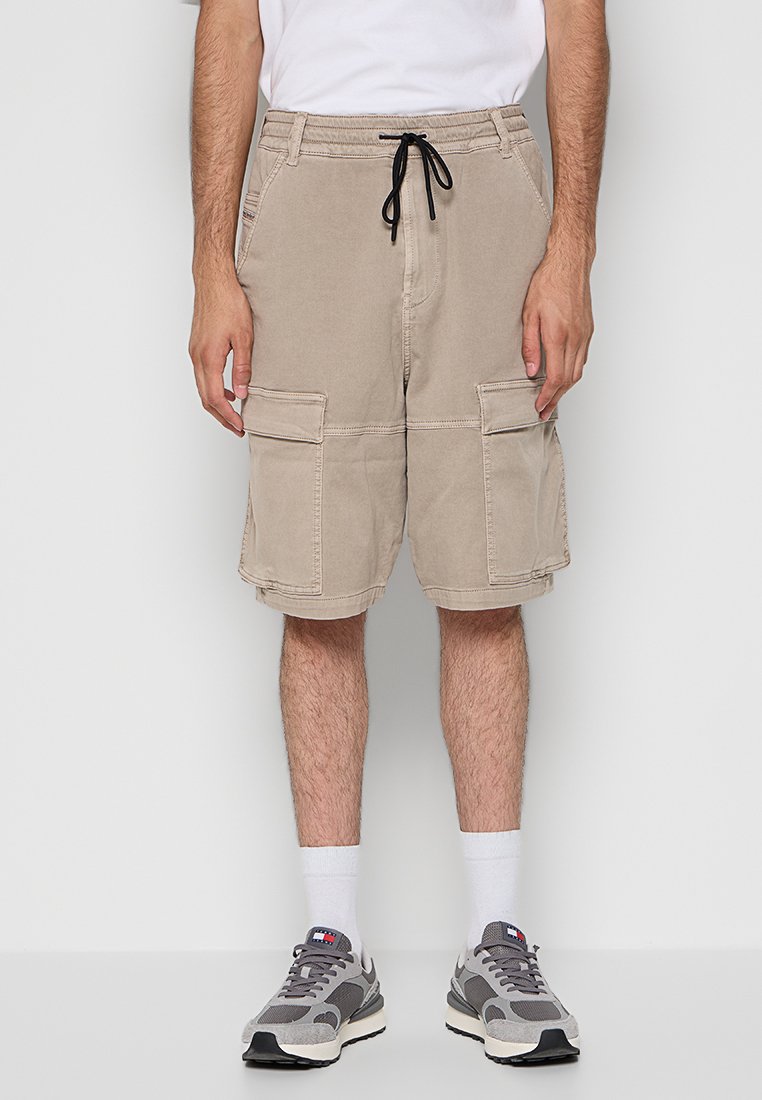 Mann in beigen Cargo-Shorts mit großen Seitentaschen, weißen Socken, grau-weißen Turnschuhen und einem weißen T-Shirt vor einem einfarbigen Hintergrund.