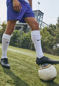 Schwarze Nike-Fußballschuhe liegen auf einem klassischen weißen Ball mit schwarzen Akzenten, getragen mit blauen Shorts und knielangen weißen Socken auf Gras.