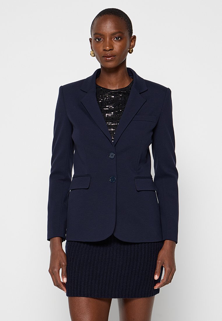 Frau trägt ein dunkelblaues Blazer über einem schwarzen Paillettenoberteil und einem passenden dunkelblauen gestrickten Minirock, mit goldenen Creolen.