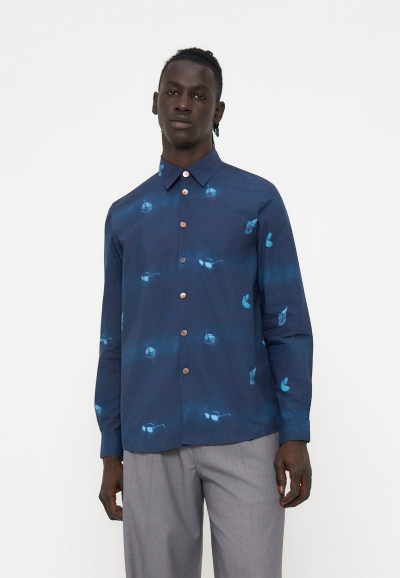 Mann trägt ein marineblaues Hemd mit Knöpfen und blauen abstrakten Mustern sowie hellgraue Hose, steht vor einem einfarbigen weißen Hintergrund.
