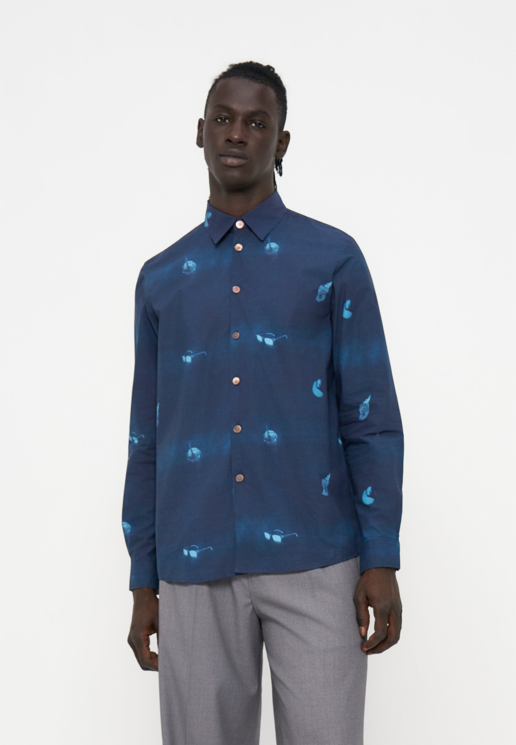 Mann trägt ein marineblaues Hemd mit Knöpfen und blauen abstrakten Mustern sowie hellgraue Hose, steht vor einem einfarbigen weißen Hintergrund.
